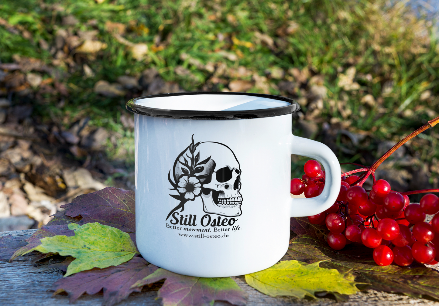 Weiße Emaille Tasse mit schwarzem Rand und Still Osteo Logo – stehend auf einem Stein in natürlicher Umgebung.